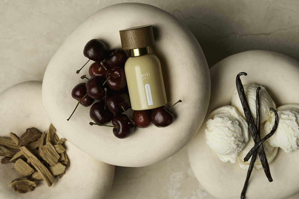 A small beige cosmetic bottle with a wooden cap and gold collar sits on a stone slab, surrounded by red cherries, vanilla ice cream with vanilla beans, and wood chips on a beige marble surface.