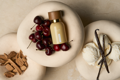 A small beige cosmetic bottle with a wooden cap and gold collar sits on a stone slab, surrounded by red cherries, vanilla ice cream with vanilla beans, and wood chips on a beige marble surface.