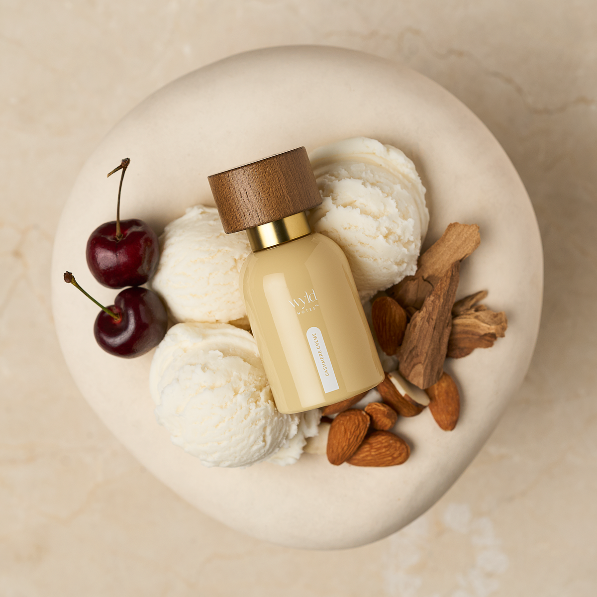 A small frosted bottle with a wooden cap and gold collar sits on a beige stone plate, surrounded by scoops of vanilla ice cream, two dark cherries, and almonds. The bottle has a small label that reads &